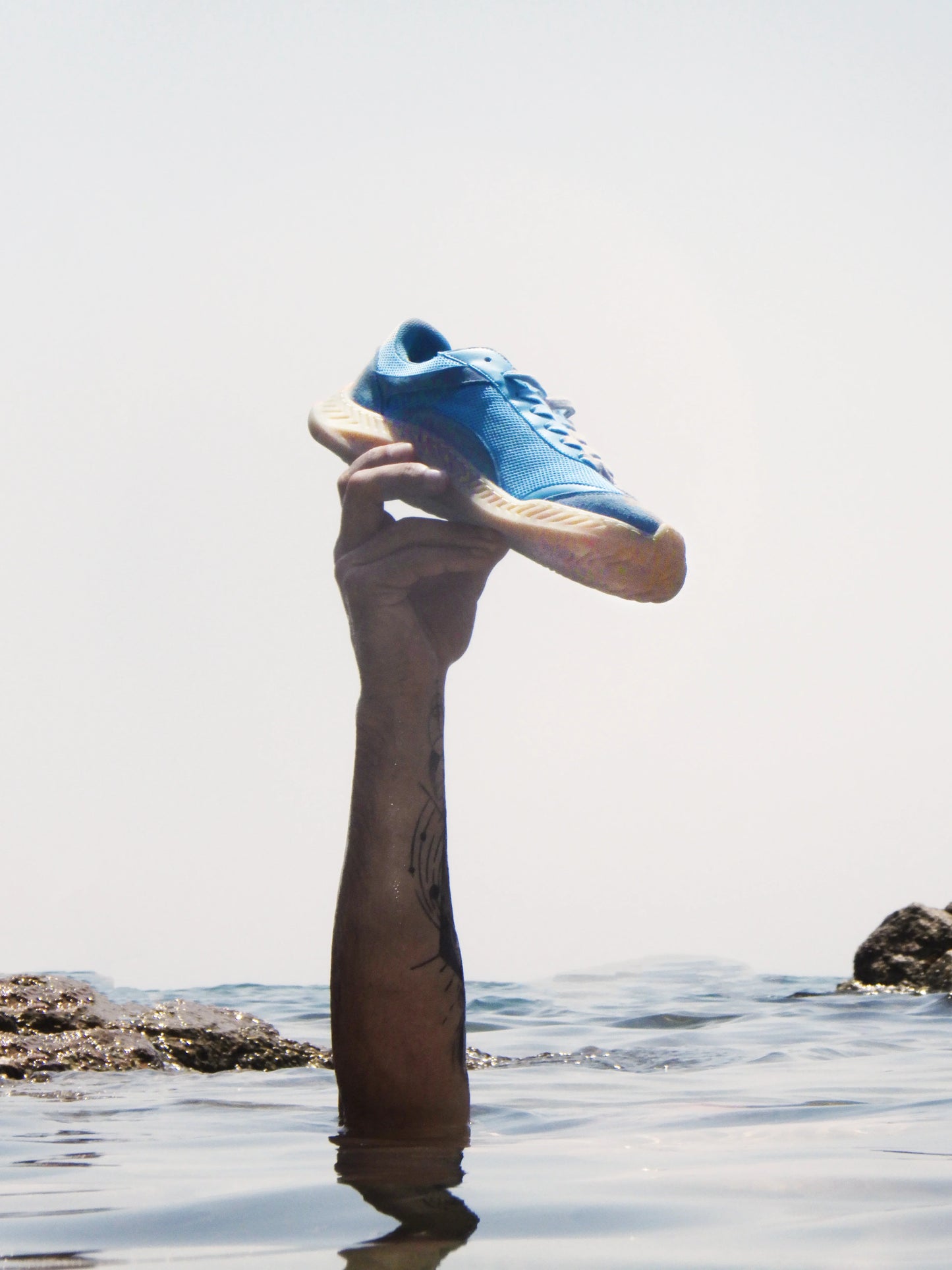 Hand holding a blue barefoot and minimal sneaker above water on a beach