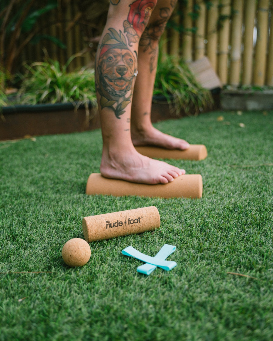 Barefoot woman balancing on The Nude Foot cork rollers, next to the cork massage ball and toe spacer band from the Foot Health & Mobility Kit