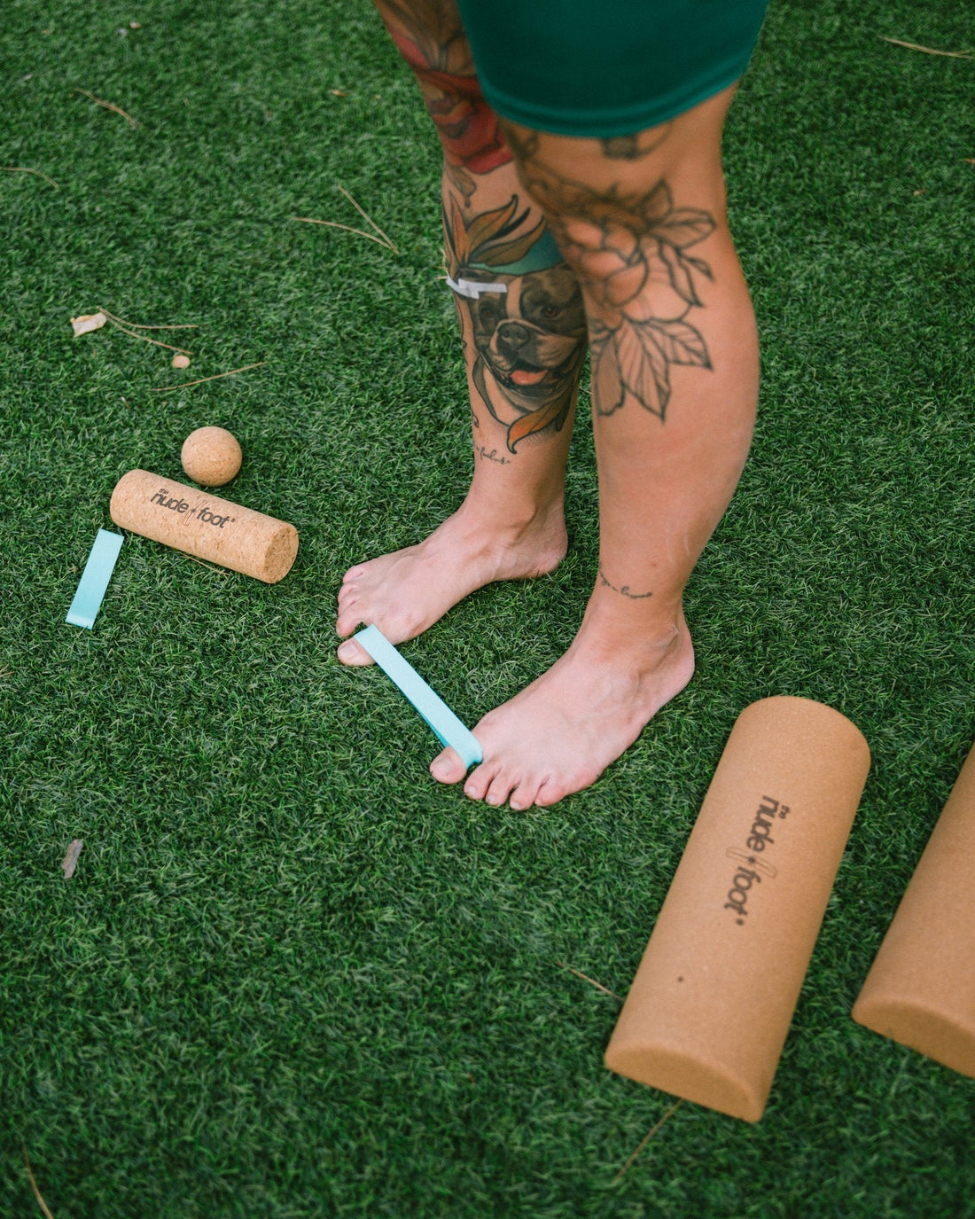Overhead view of bare feet stretched with a teal toe spacer band beside The Nude Foot cork roller and massage ball