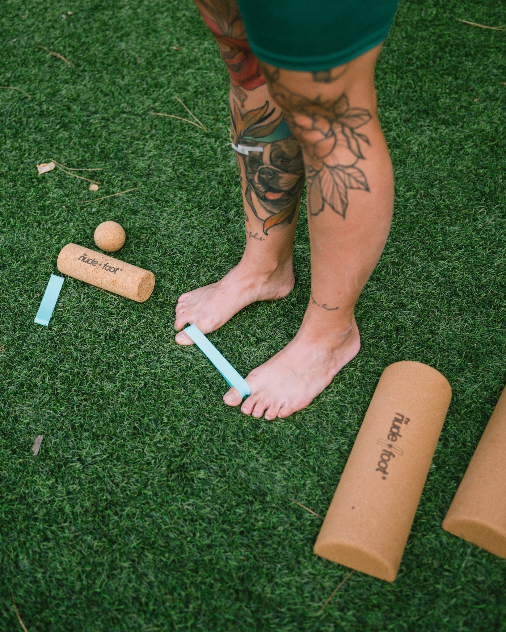 Overhead view of bare feet stretched with a teal toe spacer band beside The Nude Foot cork roller and massage ball