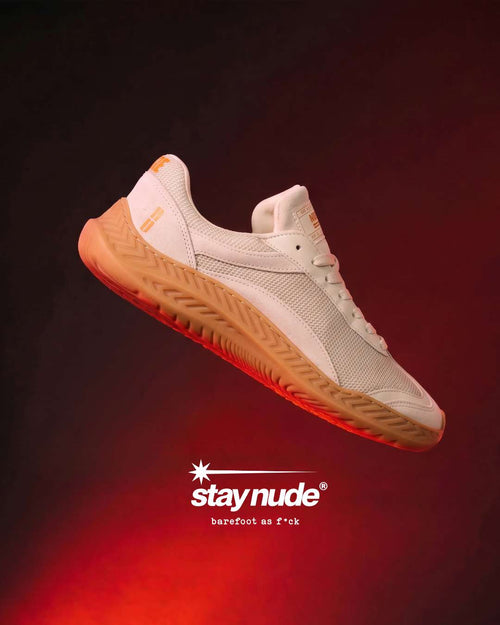 A single Stay Nude barefoot sneaker floating against a dark red and black background with dramatic lighting. The shoe is off-white with a tan gum sole and subtle orange-red glow from the lighting.