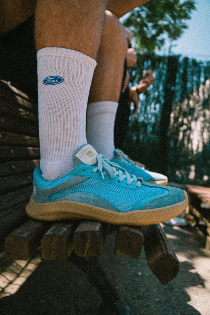 Person wearing blue barefoot and minimal sneakers and white socks with a visible brand logo, sitting on a wooden bench.