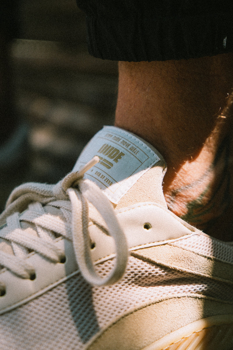 Close-up of a beige barefoot and minimal sneaker with a brand label, worn by a person with tattoos.