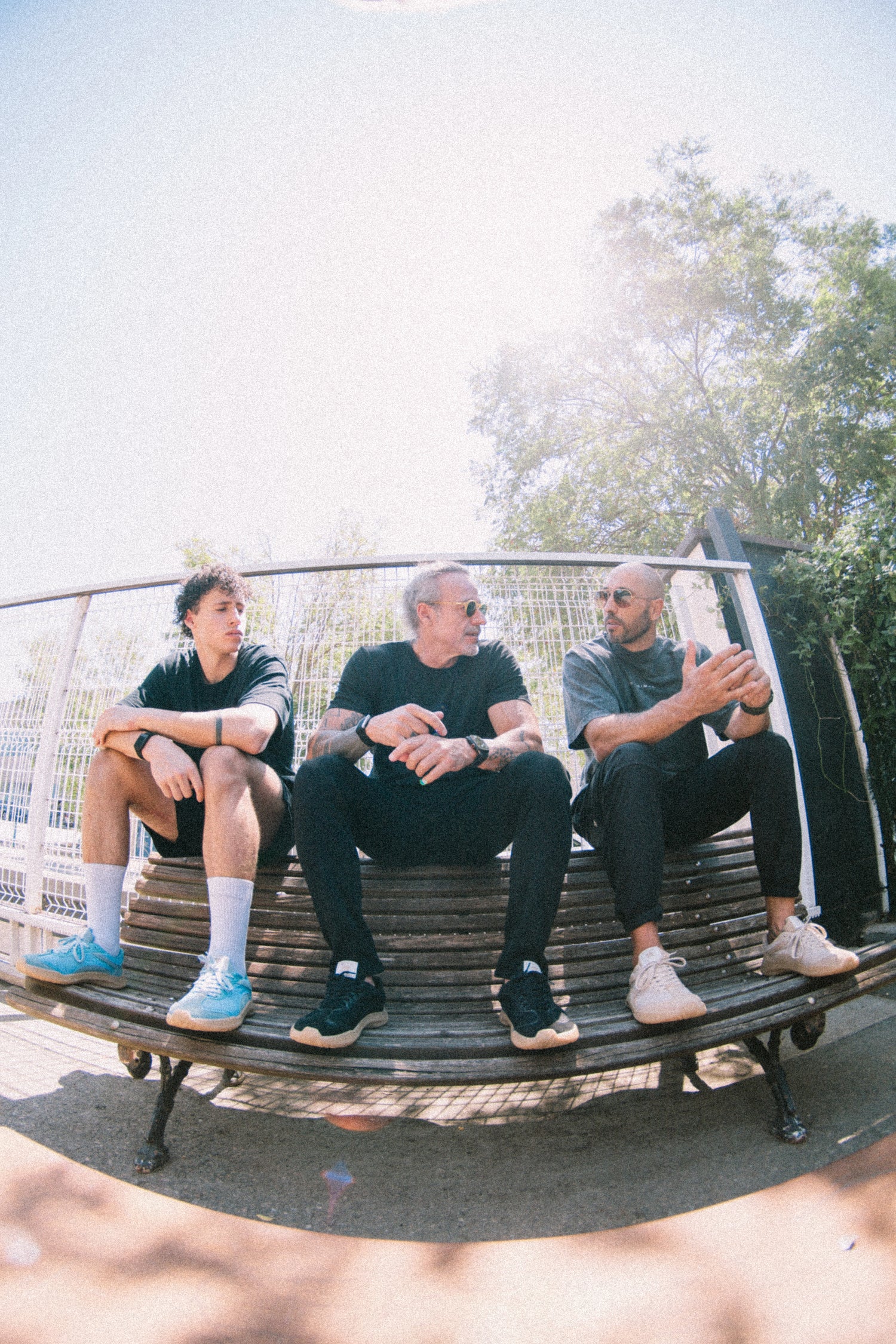 Three men sitting on a curved metal bench outdoors with barefoot and minimal  sneakers